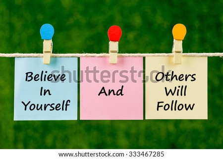 Word quotes of Believe In Yourself And Others Will Follow on sticky color papers hanging on rope against blurred green background.
