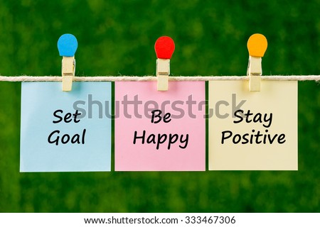 Word quotes of Set Goal, Be Happy, Stay Positive on sticky color papers hanging on rope against blurred green background.