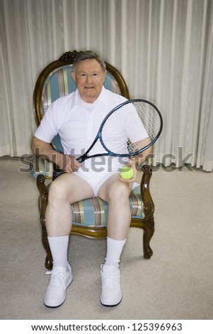 Mature man sitting in an armchair holding tennis racket and ball