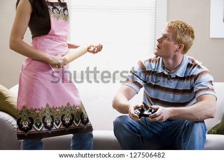 Man sitting on sofa with video game controller and woman with rolling pin in apron standing