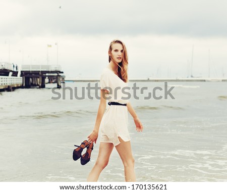 Beautiful Cute Girl Summer Walks Along The Beach Barefoot With Shoes In Hands