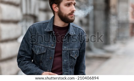 A handsome man with a beard in a jeans jacket on the street smoking
