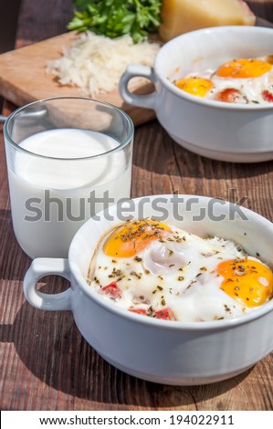eggs cocotte, baked egg with tomato,grated parmesan and glass of milk, in a white bowl, on a wooden surface, selective focus