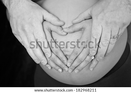 Close up of a pregnant belly in black and white; parents holding their hands in a shape of a heart