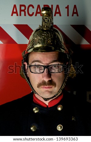 Close up self portrait of a man with a moustache wearing a brass firefighters helmet and old style woollen serge jacket with brass buttons. The man is standing in front of a fire truck from Arcadia.