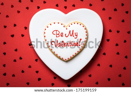 Valentine cookie in shape of heart on white plate. Be my valentine. Red paper background with confetti. Valentine\'s Day composition. Top view