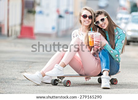 Beautiful young girls hipster girlfriends posing with a skateboard seat on skate, street fashion lifestyle in sunglasses. Keep cocktail and smiling