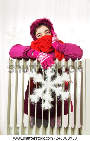 Young Girl in Warm Winter Jacket with Hood Near a Heater.