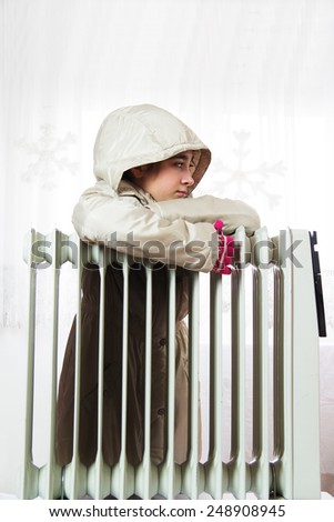Young Girl in Warm Winter Jacket with Hood Near a Heater.