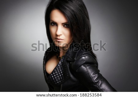 Serious rock girl, dressed up in leather jacket and hands on hips. Portrait on gray background.