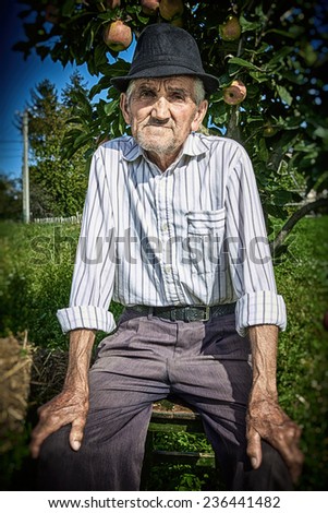 Close-up portrait of a wrinkled and expressive old farmer seated near an apple tree in the yard.