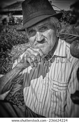 Wrinkled and expressive old farmer leaning on an apple tree in the yard. Black and white picture. Close-up.