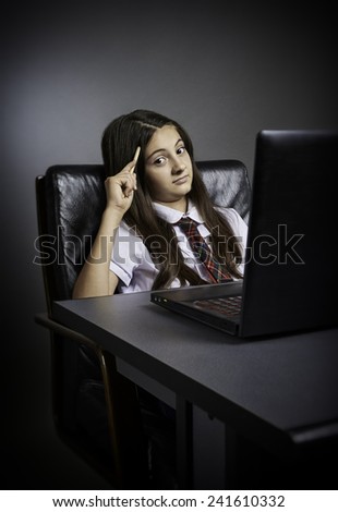 Puzzled schoolgirl sitting at desk with laptop in front and pencil in hand, looking at the camera.