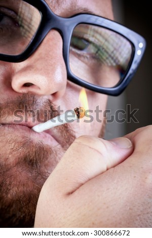 Bearded man lights a cigarette. Bespectacled man with a cigarette in his mouth and lit lighter. Portrait of a bearded with eyeglasses igniting a cigarette.