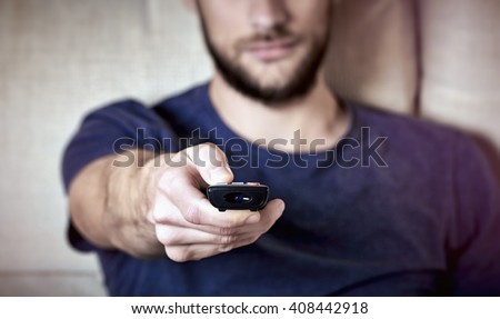 Young bearded man sitting on couch while switching TV channels. Focus on remote control.