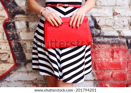 Fashion young woman with red handbag clutch in hands and striped dress near street wall closeup
