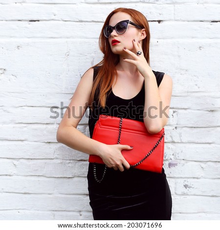closeup potrait gorgeous fashion young woman looking away and posing in black fashionable dress , sunglasses, red handbag near street white wall