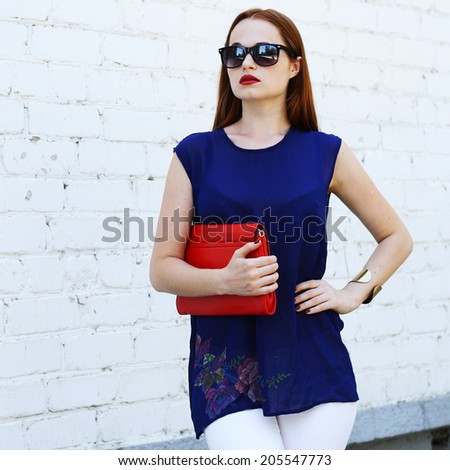 fashionable young woman wearing blue blouse and sunglasses holding handbag posing outdoors. stylish fashion portrait