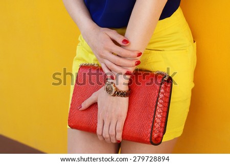 Trendy young girl in yellow short skirt and blue blouse holding red leather handbag . Outdoor shot