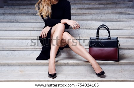 Fashion model in black dress with big leather red handbag in high heel shoes sitting on the white stair