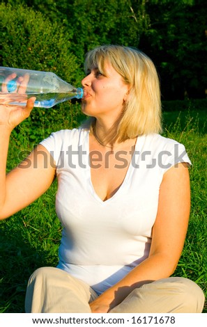 The woman-blonde drinks water from a plastic bottle against green trees
