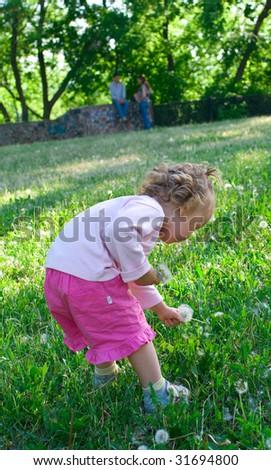 The little girl plays with flowers a lawn. Her parents talk against a trees