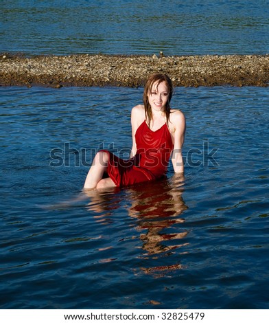 Beautiful young girl in red wet dress sits in water of the river