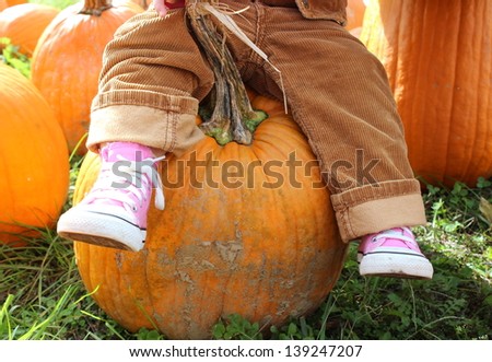 Little girl\'s legs dangling from her seat on a large orange pumpkin she singled out to take home from the local pumpkin patch.