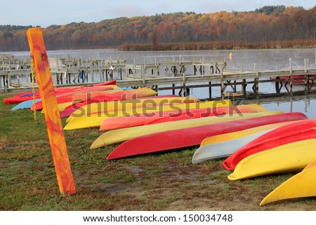 Row of colorful kayaks drying out on the shore near long wood piers at the water\'s edge, with colorful foliage beyond.