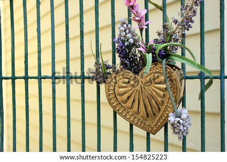 Gold metal heart with colorful floral arrangement hanging on open gate at side of building.