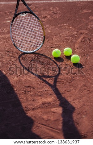 Still life of tennis balls and the shadow of a rman holding a tennis racquet on a red clay tennis court.
