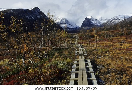 wooden path through tundra with mountain tops of singitjakka and duolbagorni in the back kebnekaise-fell, lapland, sweden