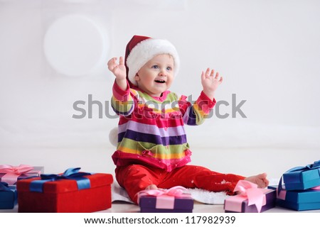 Happy baby sitting on the floor near boxes with gifts. Girl in santa hat. Christmas. The child is happy and raised his hands.