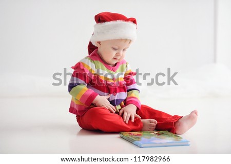 Baby sitting on the floor in a red santa hat. Child reading a book. Christmas. On the floor is a book.