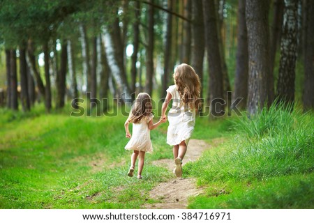 Two sisters run through the forest and holding hands. Summer sunny day and girls in light dresses. Beautiful long hair in a child.