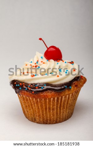 A homemade cupcake is topped with chocolate ganache, frosting, sprinkles, and a maraschino cherry