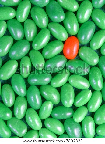 A single orange jelly bean with a large group of green jelly beans.