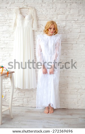 Beautiful blond bride near white brick wall inside light interior. Long wedding white dress on the wall and small table with gourmet food