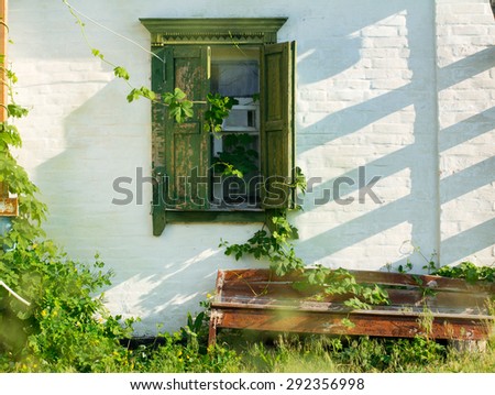 grunge white wall with green window with shutters, shadows, wild grape leaves and bench