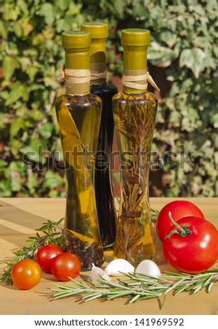 Two bottles of oil and a bottle of vinegar on a garden table with some tomatoes, garlic and rosemary.