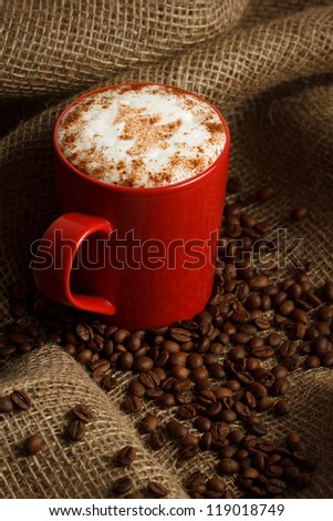 A fresh made coffee latte with Christmas tree drawing on cream. Cup standing on burlap with spread coffee beans.