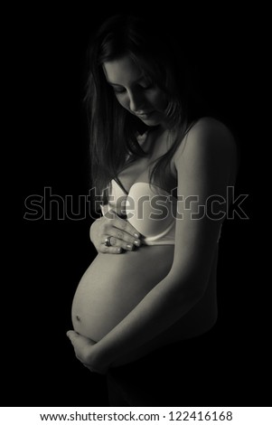 Black and white low key portrait of young pregnant woman holding her belly , isolated on black background