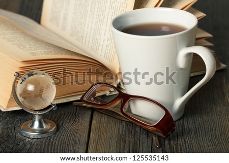 Warm tea cup on wooden table with old book, glasses and glass globe. Focused on globe.
