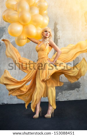 Very attractive young woman in beautiful yellow waving on a wind flow dress with yellow balloons standing on the roof.