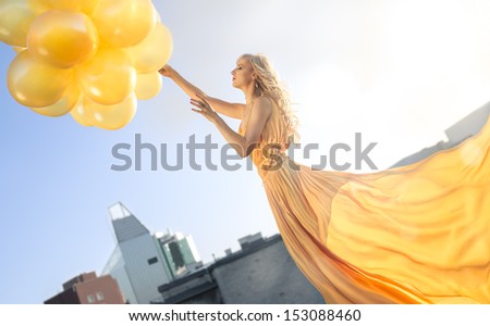 Very attractive young woman in beautiful yellow dress with yellow balloons standing on the roof.