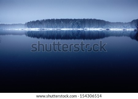 Snowfall over dark calm lake with forest on the shore