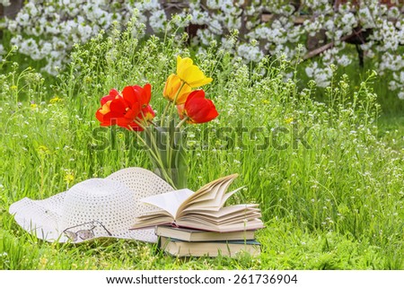 Bouquet of red tulips, books and white hat on the green grass in the spring garden against the background of cherry blossoms