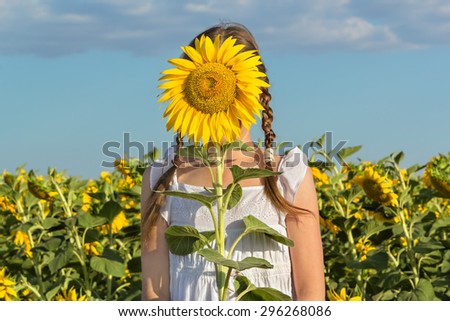Girl hiding behind flower sunflower on a background of blue sky and field with sunflowers