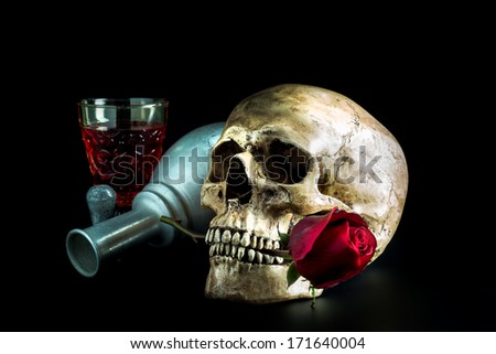 Still life with human skull with red rose bud in the mouth place beside vintage glass and bottle of wine