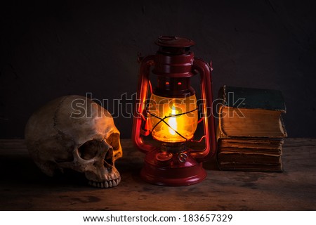 Still life with human skull and old fashioned vintage kerosene oil lantern lamp burning with a soft glow light on aged wooden floor